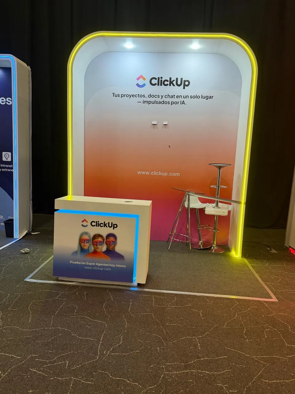 Stand de ClickUp con fondo degradado naranja y el mensaje sobre proyectos, docs y chat impulsados por IA.